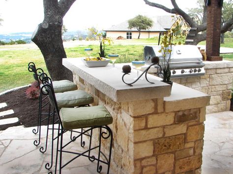 This stone paved outdoor bar features an area for enjoying drinks and entertaining guests. The space is adorned with high chairs with cushion seats and a grill unit. Diy Outdoor Bar, Outdoor Kitchen Countertops, Diy Grill, Outdoor Kitchen Bars, Outdoor Kitchen Appliances, Backyard Bar, Outdoor Bar Stools, Diy Outdoor Kitchen, Patio Bar