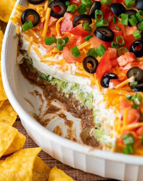 7 Layer Bean Dip with scoop taken out to show layers of beans, guacamole, sour cream and cheese. Spider Web 7 Layer Bean Dip, Black Bean Layer Dip, Halloween Nacho Dip, Easy Birthday Food Ideas, 7 Layer Bean Dip Recipe, 7 Layer Dip Easy, Layered Nacho Dip, Layered Bean Dip Recipe, Layered Taco Salad