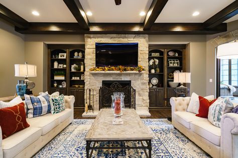 Floor-to-ceiling stone fireplace with dark stained built in cabinets and beamed ceiling. Off-white tufted sofas and stone coffeetable. The pops of red and blue give the neutral color palette life and interest. Dark Wood Built Ins, Wood Built Ins, Stone Fireplace Living Room, Tufted Sofas, White Stone Fireplaces, Edge Magazine, Rustic Family Room, Blue Bookcase, Fireplace Living Room