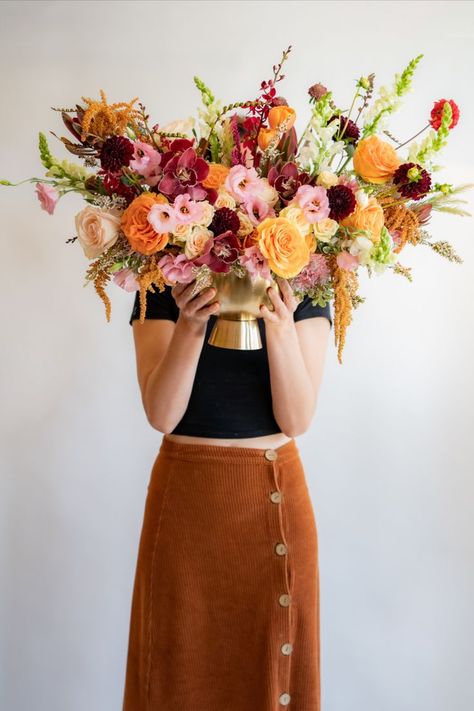 woman in black tee and brown skirt holding maroon, orange, and pink flower arrangement in gold container in front of her face Background Color Palette, Color Palette Autumn, White Delphinium, Pink And Burgundy Wedding, Palette Autumn, Red Flower Arrangements, Golden Vase, Burgundy Wedding Flowers, Burgundy Dahlia