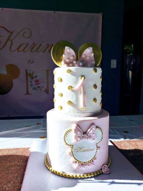 Two tier Minnie Mouse cake with gold Minnie ears on top and pink and white bow. Top tier shows child's age, and bottom tier shows child name surrounded by gold trimmed Minnie ears silhouette. Rose Gold Minnie Mouse Cake, Minnie Mouse Birthday Pink And Gold, Disney 1st Birthday Cake, Mini Mouse Cake 1st Birthday, Gold And Pink Minnie Mouse Cake, 1st Birthday Cake Minnie Mouse, Pink Minnie Cake, Pink Mini Mouse Cake, Minnie Cakes Birthday