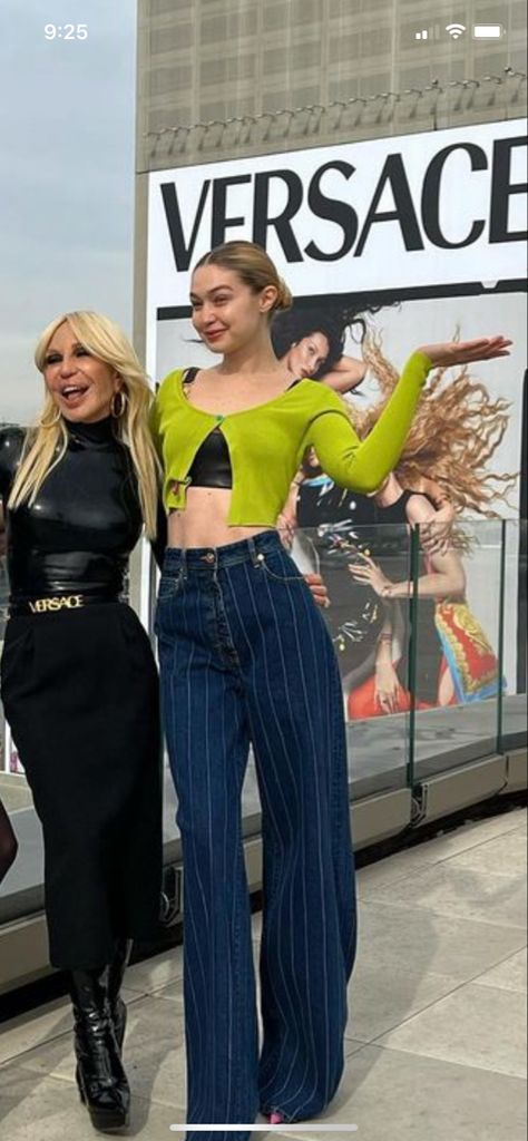 Gigi wearing a green sweater (with the length of a top) over a black top, she is also wearing dark blue jeans with white skinny vertical lines, her hair is in a middle bun and she is posing next to Donatella Versace who’s wearing a black leather o latex turtle neck-long-sleeved shirt, a black long tube skirt and a Versace black belt. They ate both in front of a Versace billboard for their most recent 2022 campaign. Gigi Hadid Outfits Casual, Gigi Hadid Off Duty, Gigi Hadid Casual, Gigi Hadid Photoshoot, Gigi Hadid Street Style, 90s Versace, Gigi Hadid Outfits, Gigi Hadid Style, Fashion Model Poses