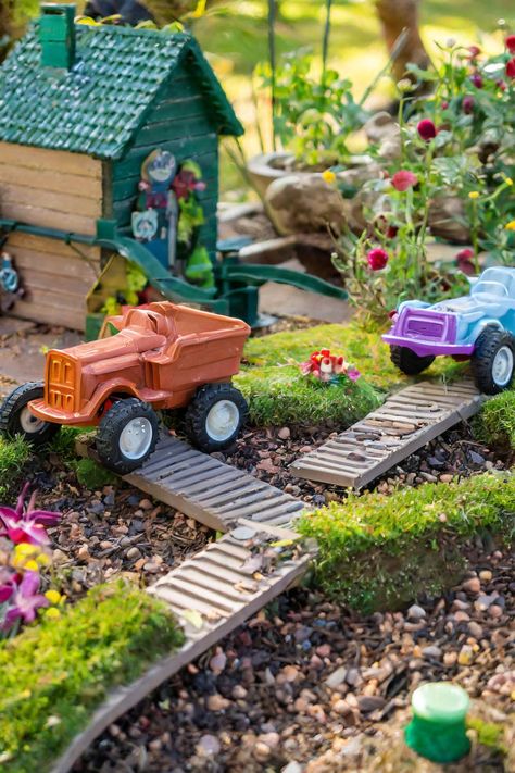 A playful fairy garden scene featuring toy monster trucks on a dirt and stone path, surrounded by flowers and a small house, perfect for gardens for children. Great inspiration for a fairy garden around a tree and tips on how to start a fairy garden with fairy tale garden ideas. Ideal for outdoor fairy garden DIY landscapes, DIY fairy garden decor, fairy house ideas DIY, and DIY outdoor fairy garden ideas. Unique Garden Ideas, Kids Fairy Garden, Unique Gardens, Fairy Gardens, Gardening For Kids, Miniature Garden, Monster Truck, Fairy Garden, Kid Friendly