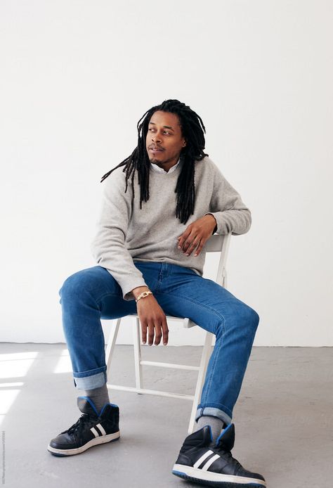 A full length portrait of an African American man with long braided hair. He has a calm confident expression and is looking off camera. He is wearing a grey sweater with a white dress collar, blue jeans and black shoes. He is casually sitting on a white chair against a white wall and there is a sun reflection the floor. Male Sitting Poses Drawing Chair, Guy Sitting On Couch Reference, Man Sitting Pose Reference Chair, Man Slouching Pose, Guy Sitting Poses, Guy Sitting In Chair, Sitting Pose Reference Chair, Man Sitting In Chair, Black Civic