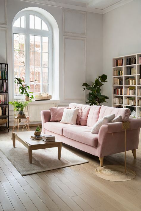 Calm Retreat • The room features an arched window that bathes it in natural light • A dusty pink sofa adds a gentle pop of color to the neutral palette • Wooden coffee table with clean lines sits atop a natural fiber rug • A tall, leafy indoor plant brings a refreshing energy to the corner • White bookshelves brimming with books suggest a love for literature • Muted tones and subtle textures throughout the room evoke a sense of peace Dusty Pink Sofa, Pink Aesthetic Home, Soft Pink Aesthetic, Natural Fiber Rug, White Bookshelves, Aesthetic Home Decor, Pink Sofa, Stylish Living Room, Natural Fiber Rugs