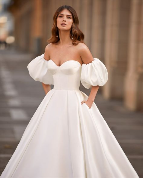 Luna Novias | This minimalist wedding dress features a tailored bodice with a voluminous pleated princess skirt and removable puffed sleeves. Tattoo Lace, Princess Style Wedding Dresses, Embroidered Wedding Gown, Beaded Wedding Gowns, Classy Prom Dresses, Minimalist Wedding Dresses, Princess Skirt, Fashion Gowns, Princess Wedding Dress