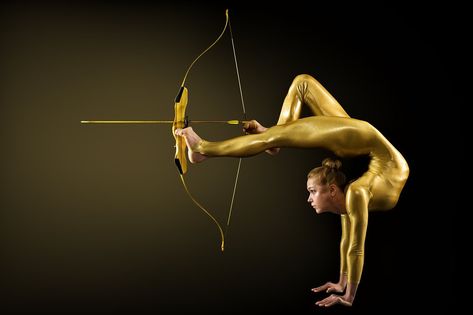 Archer Shooting by Legs with Gold Bow and Arrow. Flexible Gymnast aiming Target standing on Hand upside down. Goal Achievement Concept, Studio shot over Black background Gold Bow And Arrow, Goal Achievement, Bow Legged, Bow And Arrow, Achieving Goals, Studio Shoot, Gymnast, Upside Down, Black Background