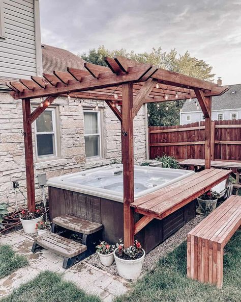 Take a soak in this brown hot tub covered by a stained wood canopy with black string lights. The canopy stands next to an adorable house made of beige stone. Make hanging out at the hot tub even easier with a wooden bar counter and wood bench. Backyard Inspiration Hot Tub, Hot Tub Ideas Backyard Pergola, Walkway To Hot Tub, Pergola Patio Hot Tub Ideas, Fenced In Hot Tub Area, Diy Hot Tub Pergola, Diy Hot Tub Table, Outdoor Patio Ideas With Hot Tub, Hot Tub Areas Outdoor