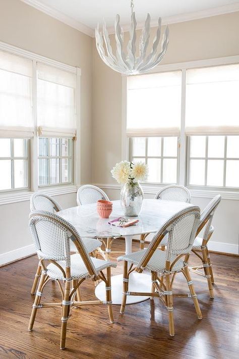 Illuminated by a glossy white leaves chandelier, an Oval Saarinen Dining Table is surrounded by Serena & Lily Riviera Side Chairs placed in front of windows dressed in white linen roman shades complementing beige painted walls in this gorgeous breakfast room. Cozy Coastal Cottage, Saarinen Dining Table, Funky Lighting, Marble Top Dining Table, Classic White Kitchen, Serena And Lily, Dining Nook, Residential Interior Design, White Leaf