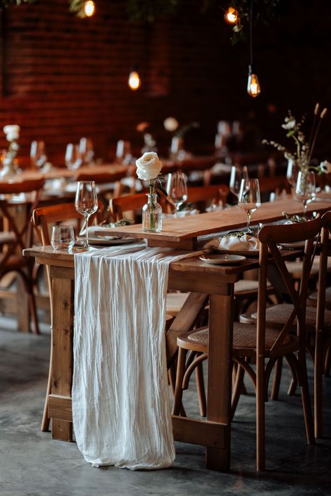 This image shows a zoom in image of a rustic dining table setup at a barn wedding. The dining furniture consists of wooden tables and chairs. On the tables are wooden table rises atop a muslin white table runner. On the table are tableware, glassware, small clear ripple glasses of flowers. Above the table are hanging boards with ivy and low hanging light bulbs with warm lighting. Farm Table Wedding, Table Risers, Trestle Tables, Runners Table, Wedding Dining, Flowers Wine, Dining Inspiration, Trestle Table, Wedding Styling