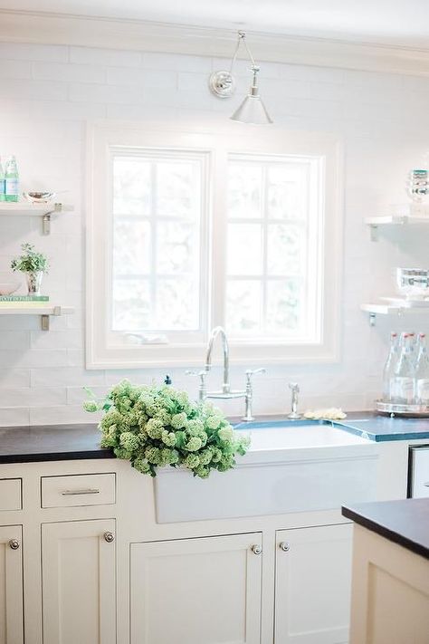 White kitchen designed with black soapstone countertops displays a white apron sink with a chrome deck mount faucet under a kitchen window surrounded by white subway tile and a chrome task sconce. Chrome Kitchen Hardware, Black Soapstone Countertops, White Apron Sink, Black Soapstone, Bathroom Feature Wall, Top Kitchen Cabinets, Cheap Kitchen Remodel, White Shaker Kitchen, Soapstone Countertops