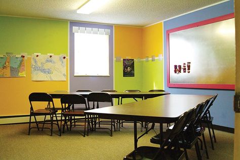 A classroom with two sets of tables and chairs. The walls are painted vibrant shades of green, orange, and blue. There is a tin magnet board on one wall, and posters on the other. School Wall Decoration Ideas, Classroom Walls Paint, Sunday School Room Decor, Inexpensive Decorating Ideas, Sunday School Classroom Decor, Church Nursery Decor, Kids Church Rooms, School Wall Decoration, Sunday School Decorations