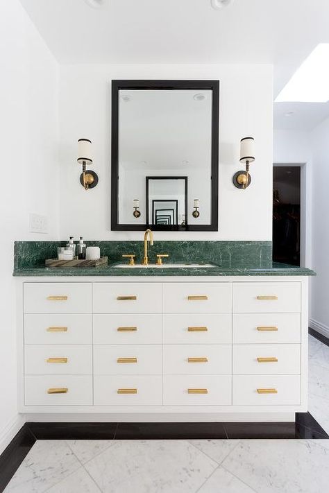 Marble floor tiles framed by black tiles lead to a white washstand fitted with brass hardware and a green marble countertop lined with a green marble backsplash. Green Marble Bathroom, Marble Countertops Bathroom, Marble Kitchen Counters, Black Marble Bathroom, Green Countertops, Bathroom Paneling, Marble Countertops Kitchen, Farmhouse Backsplash, Grey Backsplash