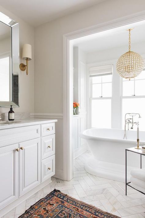 A gold French chandelier lights a rolltop freestanding bathtub matched with a polished nickel tub filler and placed in a nook on marble herringbone pattern floor tiles beneath windows and in front of white built-in cabinets. Neutral Bathroom Paint, Neutral Bathroom Paint Colors, Vanity Diy, Neutral Bathroom Decor, Bathroom Vintage, New Classic Furniture, Neutral Bathroom, Bathroom Paint Colors, In Bathroom