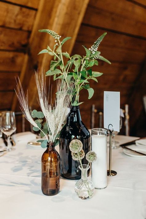 a boho wedding centerpiece of apothecary bottles, a candle, wheat and greenery for a simple look - Weddingomania Rustic Mountain Wedding Centerpieces, Greenery Vase Centerpiece, Zero Waste Wedding Decorations, Wedding Centerpieces Bottles, Boho Mountain Wedding Decor, Pottery Centerpieces Wedding, Colorado Wedding Decor, Mountain Centerpieces Wedding, Mountain Cabin Wedding