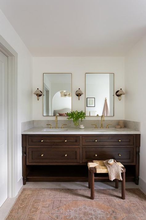 Bathroom features a brown wooden dual washstand with gray marble countertop, built in towel shelf, aged brass knobs and brass faucets under brass mirrors lit by sconces with gray pleated shades and a pink and orange vintage rug. Dark Wood Vanity, Dark Brown Bathroom, Walnut Vanity, Dark Wood Bathroom, Wood Bathroom Cabinets, Dark Bathrooms, Bathroom Vanity Designs, Bathroom Features, Dark Wood Cabinets