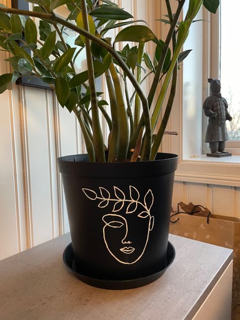 Black flowerpot with a painted one-line face with leaves Painting On Plastic Pots, Flowerpot Paintings, Flowerpot Painted, Flower Vase Designs Painted, Diy Painted Flower Pots Design, Easy Pot Painting Ideas, Black Pot Painting Ideas, Flower Vase Design, Diy Room Decor For Teens