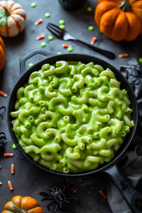 Green mac and cheese served in a black cast iron skillet, surrounded by Halloween decorations like pumpkins and spider toys. The bright green color of the pasta contrasts with the dark and festive setting. Green Mac N Cheese Halloween, Halloween Max And Cheese, Green Mac And Cheese Halloween, Spooky Mac And Cheese, Mac And Cheese Halloween, Halloween Mac And Cheese, Green Mac And Cheese, Halloween Main Dish, Halloween Party Menu