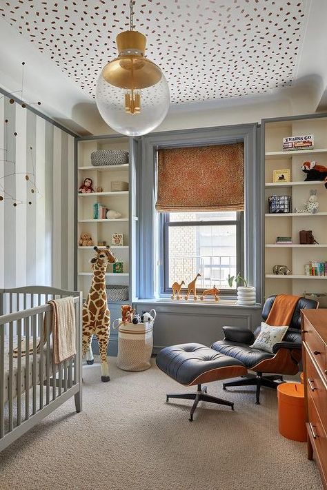 Boy's nursery features an Eames management chair and ottoman, a gray crib, a tall stuffed giraffe and built in shelves that surround a window under gold confetti ceiling wallpaper. Kids Wallpaper On Ceiling, Desi Perkins Nursery, Wallpaper Ceiling Boys Room, Nursery Wallpaper On Ceiling, Wallpaper For Built Ins, Post Modern Nursery, Built In Crib, Modern Eclectic Nursery, Nursery Ceiling Wallpaper