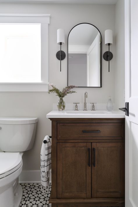 Natural elements, like a wood vanity and bud vase with greenery, can go a long way in a small powder bathroom. To add a punch of personality, our designers love incorporating a patterned floor tile to spice things up in the small space! With neutral walls and soft metal finishes for contrast, the play on pattern can really keep things interesting! Black And White Tile Floor Bathroom Wood Vanity, Bathroom Vanity Dark Wood, Bathroom Remodel Black Vanity, Half Bathroom Floor Ideas, White And Brown Bathroom, Dark Wood Bathroom Vanity, Bathroom With White Vanity, Vase With Greenery, Small Powder Bathroom