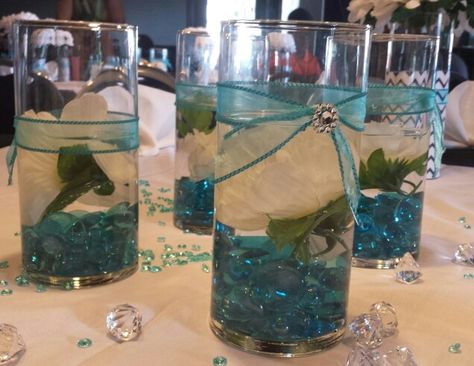 Teal table centerpiece.  Perfect for any occasion.  Floating white silk flowers  add a special touch along with teal crystals/gems. Simple and inexpensive.  Vases  and larger teal gems purchased from Dollar Tree. Ribbon and smaller teal crystals from local craft store. Teal And Gold Table Decorations, Light Teal Wedding, Teal Centerpieces, White Wedding Table Decor, Gold Table Decorations, Night To Shine, Floating Centerpieces, Teal Table, Teal Decor