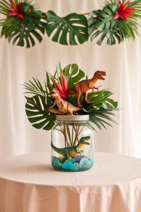 A jar on a table holds toy dinosaurs, perfect as a baby shower centerpiece, surrounded by tropical leaves and vibrant red flowers. Above, a decorative garland of large green leaves and red blooms elegantly hangs against a white curtain backdrop. Unique Centerpiece Ideas, Dinosaur Party Centerpiece, Baby Shower Centerpiece Ideas, Storybook Theme, Dinosaur Baby Shower, Rubber Ducks, Unique Centerpieces, Budget Friendly Decor, Shower Centerpieces