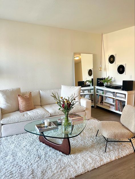 Apartment living room with white couch and throw pillows, a Noguchi mid century modern glass coffee table with coffee table books, a candle, and flowers. A credenza with a record player and speakers, styled with books, records, and flowers, during sunset Vogue Room Ideas, Coffeetablebooks Deco, Girly Apartment Living Room, Cool Girl Apartment, College Living Room Apartment, Bachelorette Pad Apartment, Trendy Apartment Decor, Apartment Deco, Girl Apartment