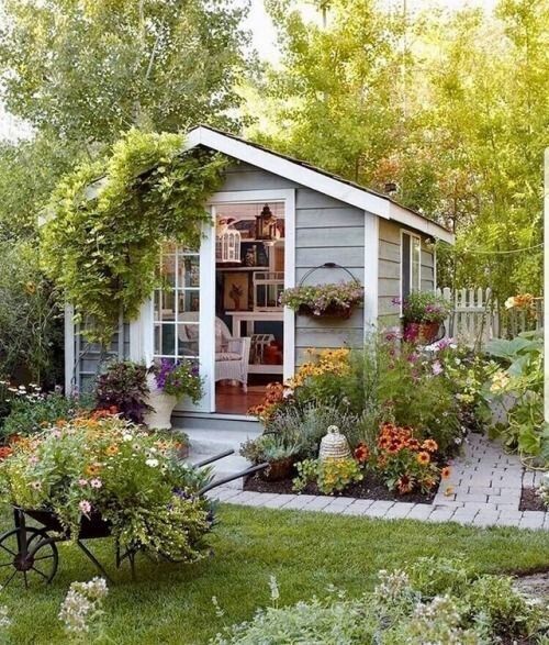 a garden shed with lots of flowers and plants in the front yard, next to a wooden bench