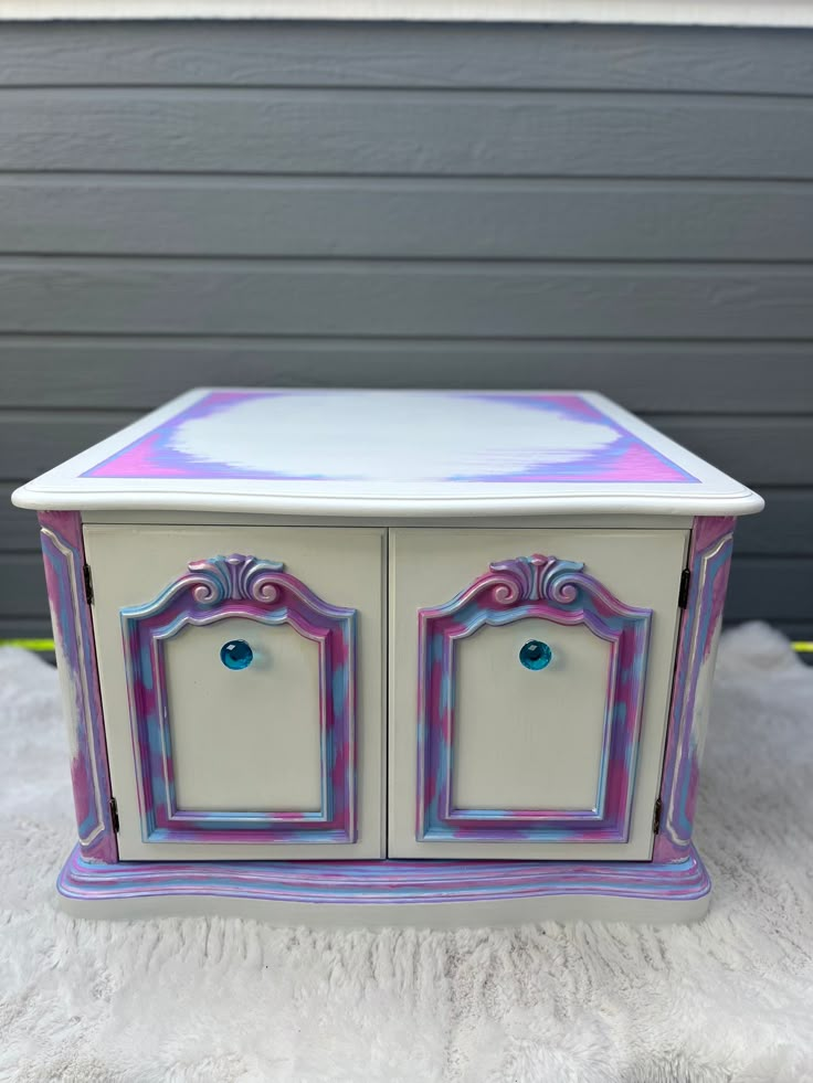 a white and purple cabinet with two doors on the front, sitting on a bed