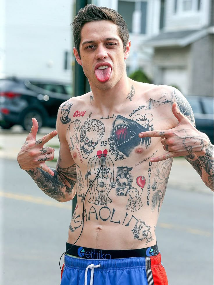 a shirtless man with his tongue out and tattoos on his chest is making the peace sign