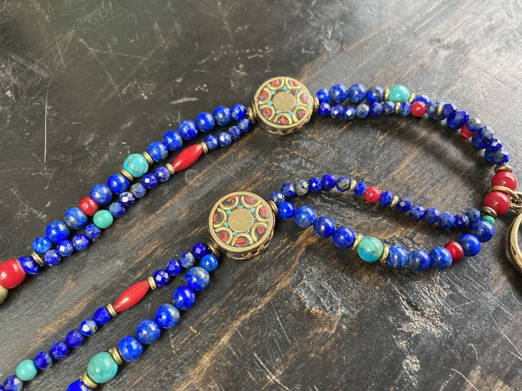 Featuring stones that are associated with wisdom, insight, spirituality, and communication, this is made with gorgeous deep royal blue to navy blue grade A Lapis in various shapes and sizes, which contrast strikingly with green semi precious turquoise, rich red Czech glass and white-heart beads, and brass spacers. The two strands gather at Tibetan brass 15mm coin beads inlaid with red coral and turquoise howlite as well as other brass beads. A 53x37mm plated antique gold large tribal style medal Spiritual Blue Beaded Necklaces For Festival, Spiritual Blue Beaded Necklaces For Healing, Blue Lapis Lazuli Jewelry For Festivals, Blue Spiritual Beaded Necklace For Festival, Spiritual Blue Necklaces With Polished Beads, Blue Necklace With Colorful Beads For Meditation, Spiritual Blue Necklaces With Natural Stones, Blue Spiritual Necklace With Gemstone Beads, Blue Spiritual Gemstone Bead Necklace