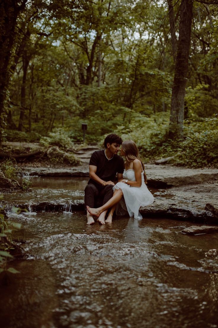 a man and woman sitting on rocks in the middle of a stream with trees around them