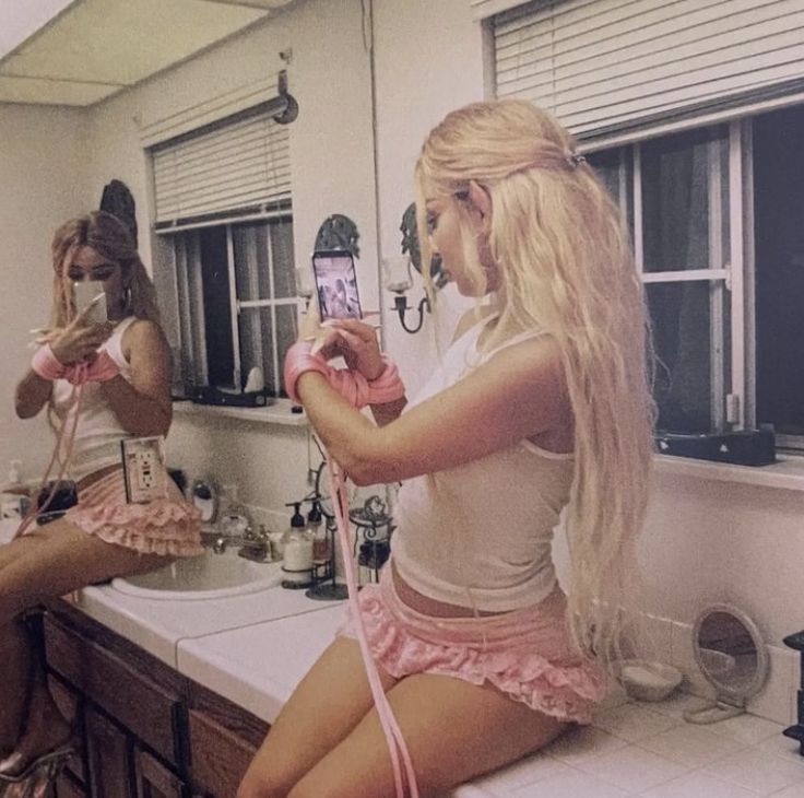 two women sitting on a counter in a bathroom taking pictures with their cell phones and hair dryers