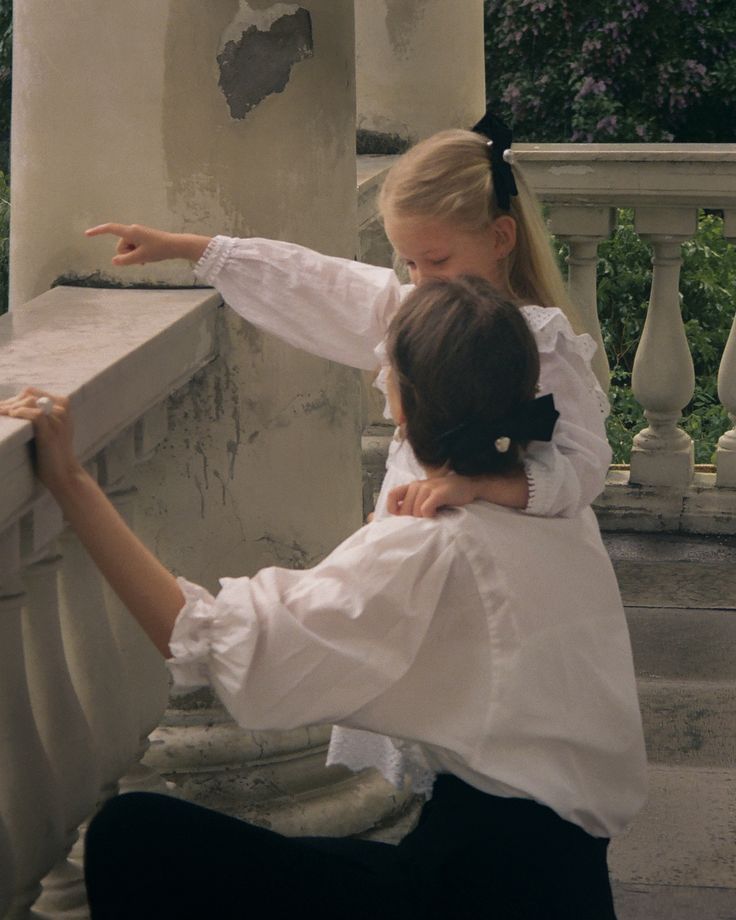 two girls in white shirts and black pants leaning against a stone wall with their arms around each other