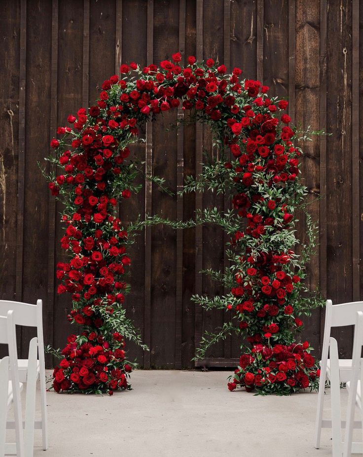 an outdoor ceremony with white chairs and red flowers on the arch, set up in front of a wooden wall