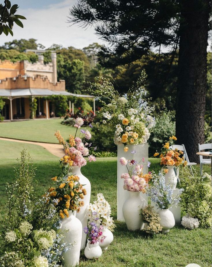 several white vases with flowers in them sitting on the grass near some bushes and trees