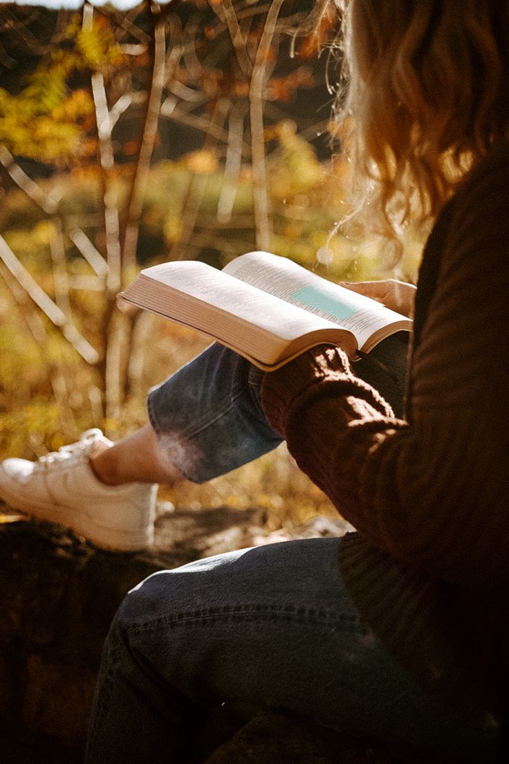 a person sitting down reading a book in the fall sun with their feet propped up