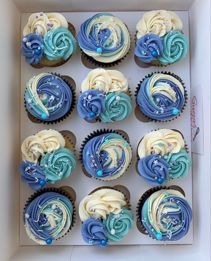 twelve cupcakes with blue and white frosting in a box on a table