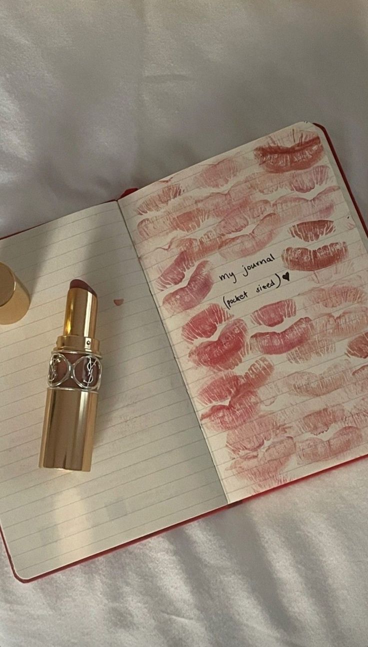 an open notebook with lipstick on it next to a bottle of perfume and a pen