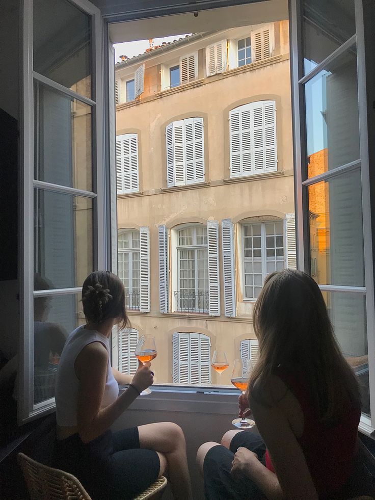 two women sitting in front of a window drinking wine and looking out at the city