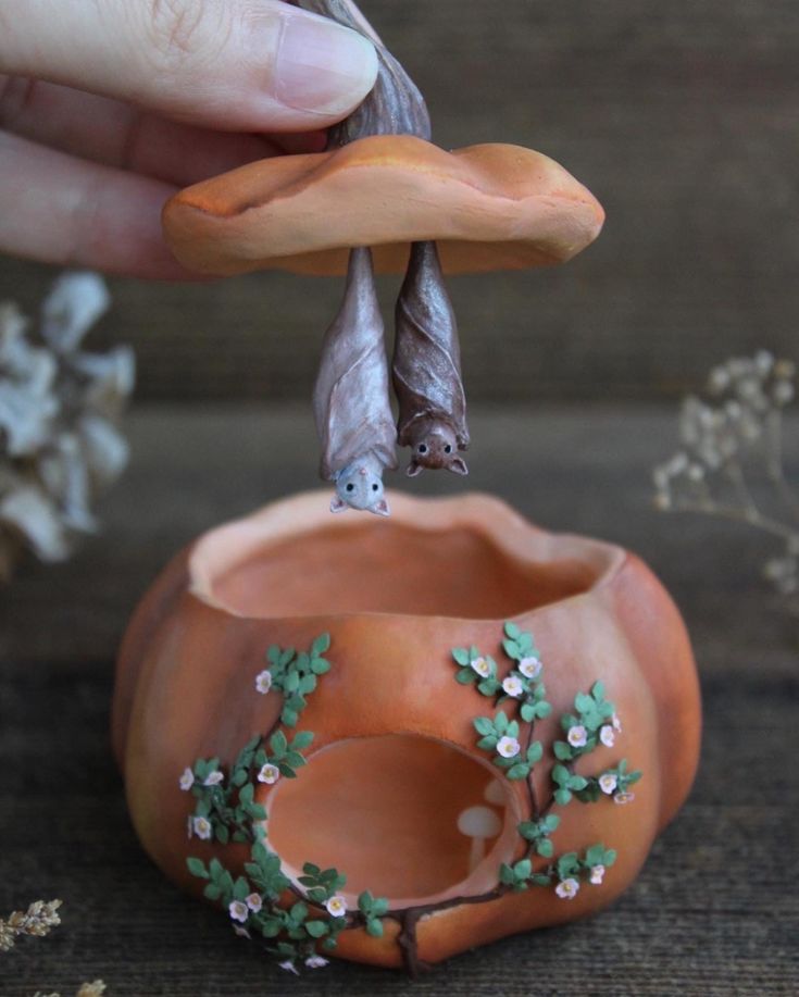 a hand is holding an orange vase with two bats on it and flowers in the background