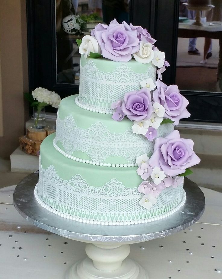 a three tiered cake with purple flowers on the top and white lace around the edges