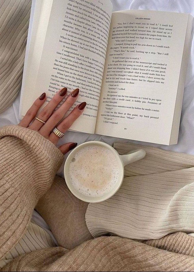 a woman laying in bed reading a book and holding a cup of coffee