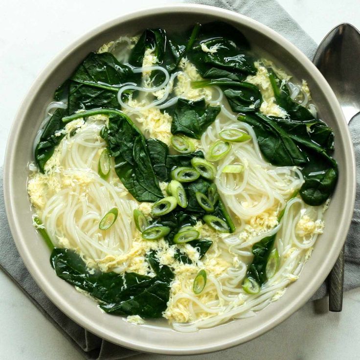 a bowl filled with noodles and spinach on top of a white table cloth next to a spoon