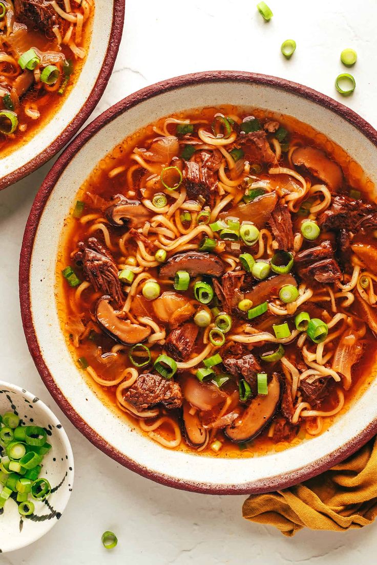 two bowls of beef noodle soup on a white surface with green onions and peas