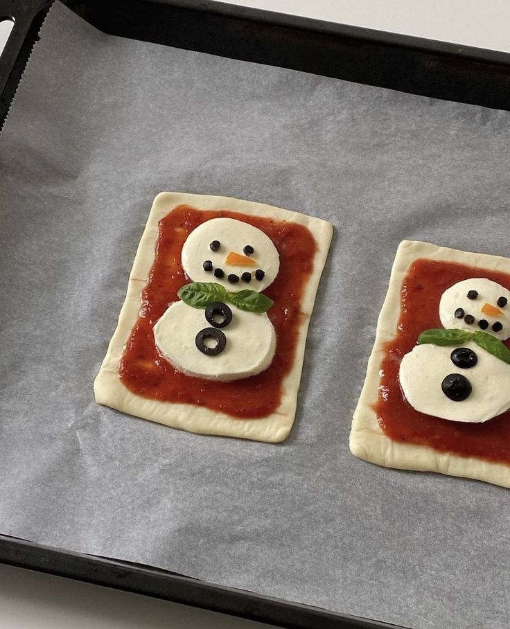 two snowmen made out of dough sitting on top of a baking sheet with tomato sauce and olives