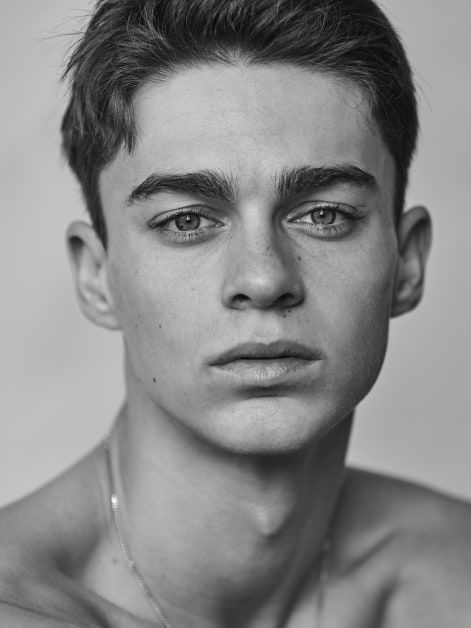 black and white photograph of a young man with necklace on his neck looking at the camera