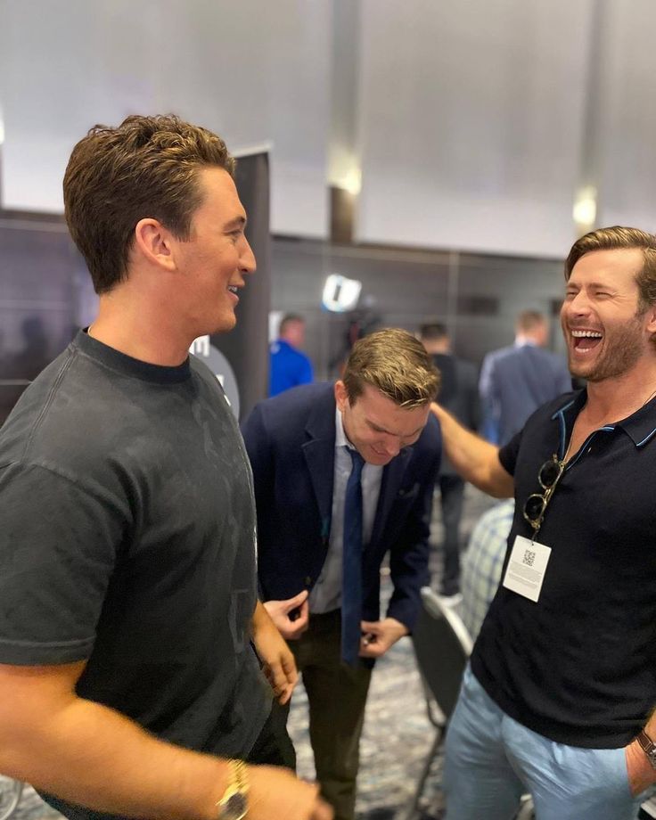 two men laughing and standing next to each other in an office building with other people