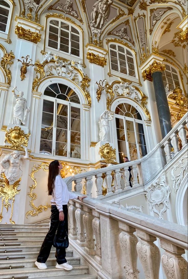 a woman walking up the stairs in an ornate white and gold building with large windows