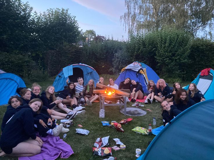 a group of people sitting around a campfire in the grass with tents behind them